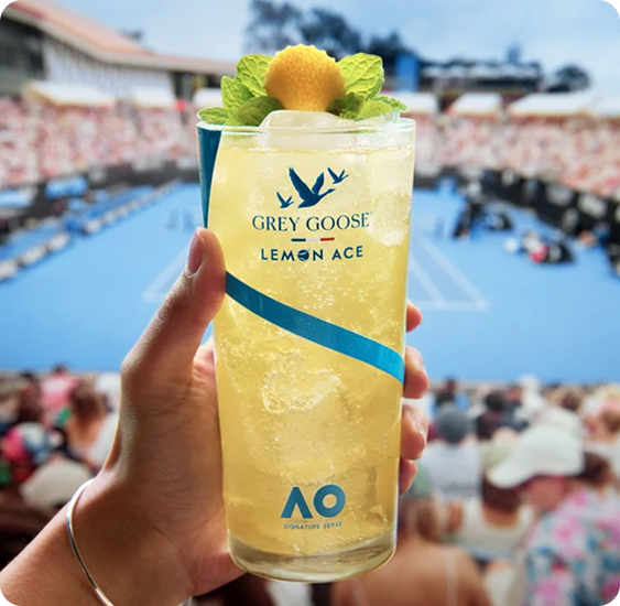 A close-up of a hand holding a tall Grey Goose Lemon Ace cocktail filled with crushed ice, garnished with fresh mint and a lemon peel, in a branded cup. In the background, a sunlit tennis stadium is filled with spectators watching a match on a bright blue court at the Australian Open, creating a lively summer atmosphere.