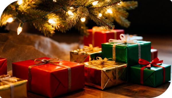 Close-up of assorted Christmas presents wrapped in red, gold, and green paper placed under a Christmas tree with warm lights.