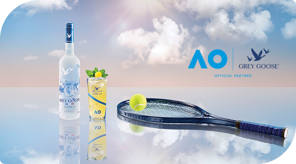 Bottle of Grey Goose vodka and a Grey Goose Lemon Ace cocktail with two lemons arranged on a wooden tray in the shape of a tennis racket on a blue tennis court with two tennis balls and the Australian Open and Grey Goose logos in the background.