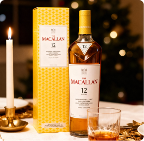 The Macallan Color Collection 12 Years Old single malt whisky bottle with its gold box and a glass of whisky over a table at dinner time with warm lights, a white candle and a Christmas tree on the background