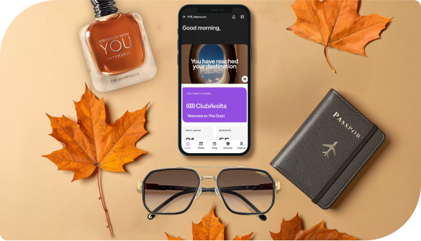 Flat lay of autumn-themed items including 'Stronger with you Intensely' by Emporio Armani fragrance, the Club Avolta app open on a cell phone screen, sunglasses, a passport case, and several orange maple leaves.